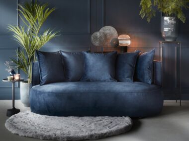 أريكة مخملية بمقعدين باللون الأزرق الداكن بأشكال دائرية ووسائد ظهر متطابقة. مصممة بسجادة مستديرة ونباتات.