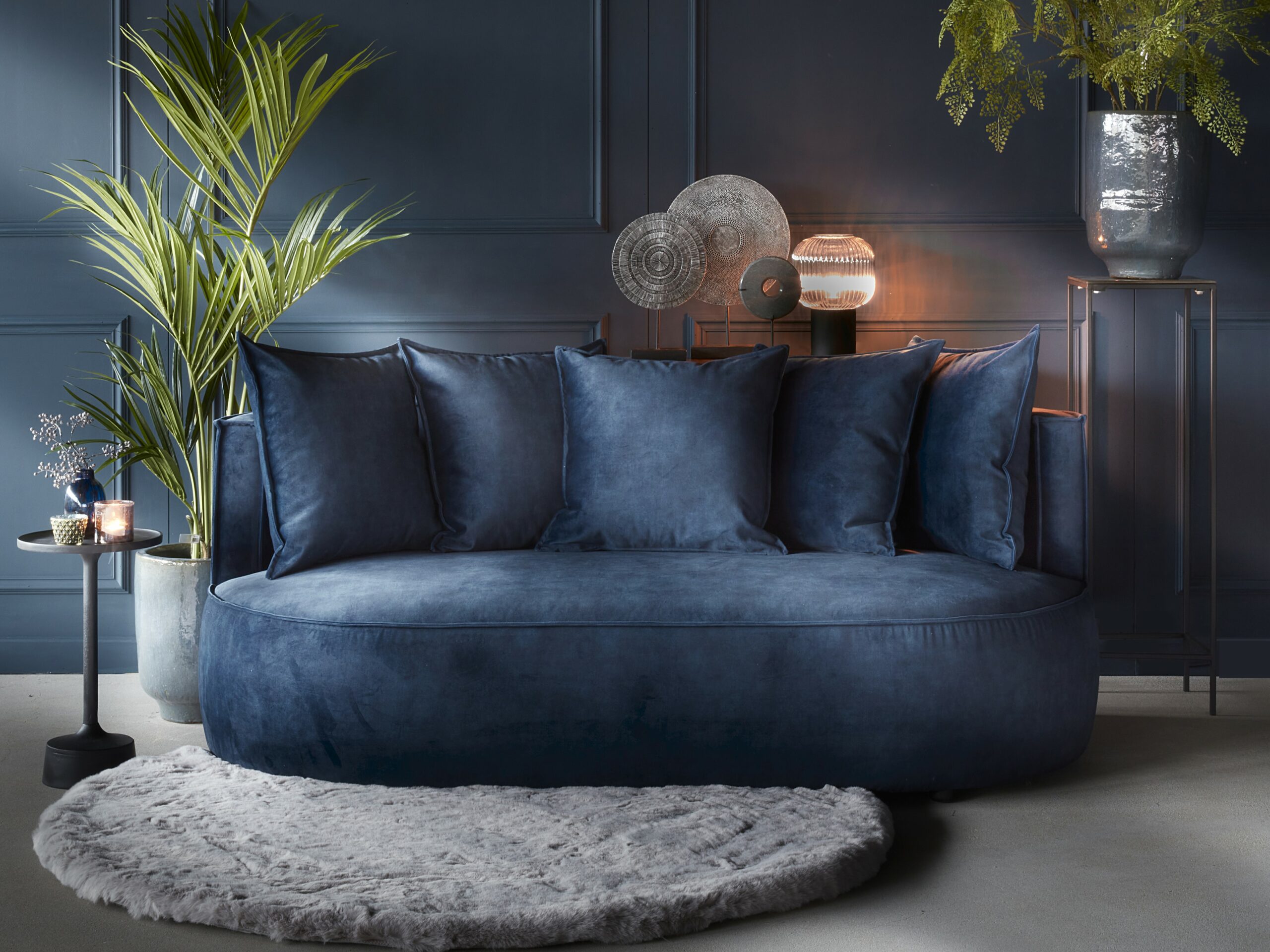 Sametová tmavě modrá dvoumístná pohovka s kulatými tvary a odpovídajícími opěradly. Stylizované s kulatým kobercem a rostlinami.