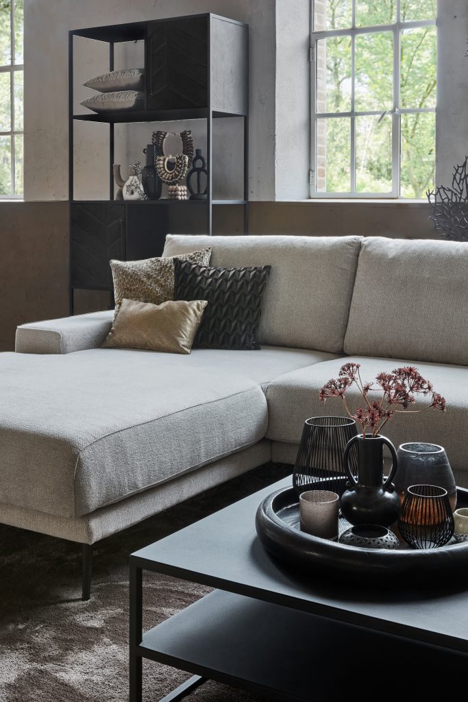 Canapé d'angle Rosalie en tissu beige. Décoré avec des meubles en métal noir et un grand tapis. Photo détail avec accessoires et coussins décoratifs.